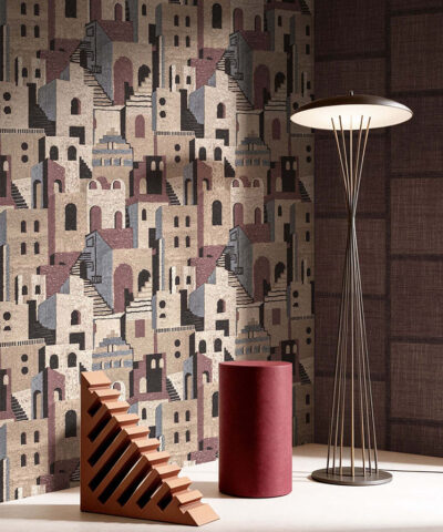 Carta da parati GAIA con villaggio stilizzato nei toni beige, bordeaux e antracite, in ambientazione elegante con lampada da terra e oggetti decorativi